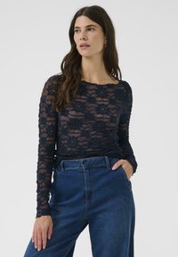 Vrouw met lang bruin haar, gekleed in een marineblauw bloemetjes kant top met lange mouwen en hoog-taille blauwe wijde jeans, staand met één hand in de zak.