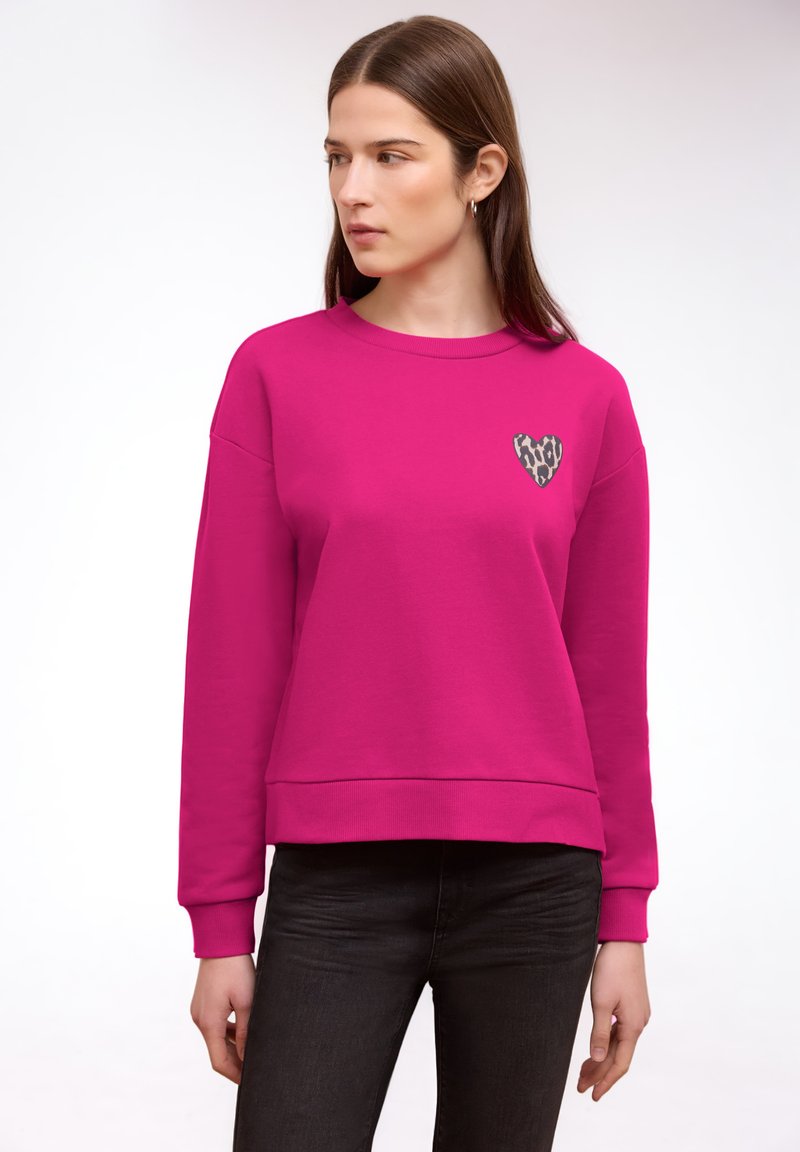 Femme portant un sweat-shirt rose vif avec un cœur imprimé léopard sur la poitrine, associé à un jean noir, regardant sur le côté.