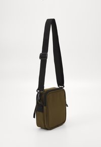 Malá olivově zelená crossbody taška s černým nastavitelným popruhem a zipovou hlavní přihrádkou na bílém pozadí.