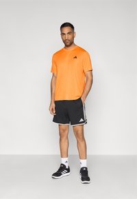 Orange athletic t-shirt with black logo, black shorts with white stripes and logo, black shoes with white accents, standing against a light background.