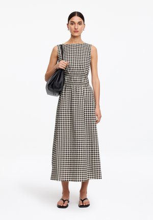 Mujer con un vestido sin mangas de cuadros vichy en blanco y negro con la cintura ceñida, llevando un bolso negro al hombro y sandalias negras.