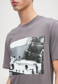 Grå bomulds t-shirt med en sort-hvid grafisk tryk af en badekar scene med planter og dekorative genstande. Rund halsudskæring, korte ærmer.