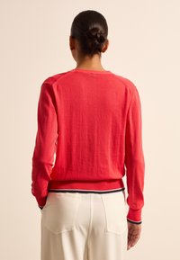 Roter Strickpulli mit langen Ärmeln, gerippten Bündchen und Saum, der am unteren Rand mit einem navyblauen und weißen Streifenakzent versehen ist.