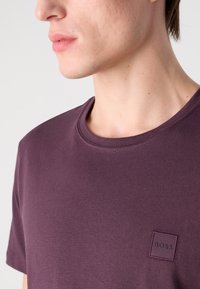 Tricou cu guler rotund de culoare mov, confecționat dintr-un material moale, având un mic etichetă cu logo în formă de pătrat pe partea stângă a pieptului. Design simplu și minimalist.