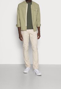 Lys grøn jakke med krave og forlommer over en oliven grøn t-shirt, kombineret med beige bukser og hvide sneakers.