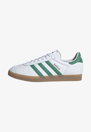 Baskets blanches Adidas Gazelle avec des rayures vertes, semelle en gomme, lacets et texte "GAZELLE" sur le côté.