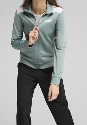 Femme portant une veste zippée Puma vert clair avec des panneaux blancs sur les épaules et un pantalon noir sur un fond uni.