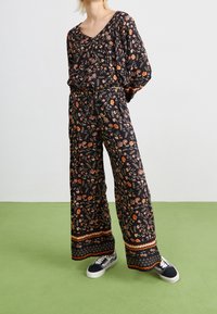 Combinaison noire à motif floral avec un décolleté en V, des manches larges et une taille à cordon. Présente des accents orange et blancs au niveau de l'ourlet. Associée à des baskets foncées.