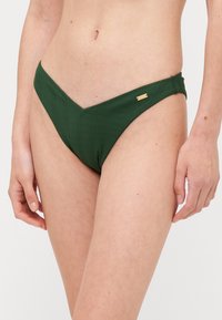 Mörkgrön bikiniunderdel med v-formad framsida, texturerad ribbad design och diskret detaljer i guldfärg på sidan.