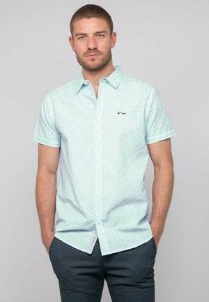 Homme avec une barbe courte portant une chemise bleu clair à manches courtes boutonnée et un pantalon sombre à motifs, debout les mains dans les poches.