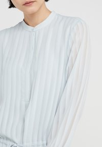 Himmelblaue Bluse mit vertikalen Streifen, Knopfleiste am Hals und langen, durchsichtigen Ärmeln. Der Stoff wirkt glatt mit einer leichten Textur.