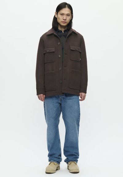 Chaqueta de lana marrón con grandes bolsillos frontales, cierre de botones y cuello puntiagudo, combinada con pantalones vaqueros holgados azules y zapatos beige.