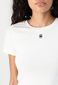 T-shirt bianco in cotone con colletto tondo e maniche corte. Caratterizzato da un piccolo logo nero "RAW" nella zona del petto. Texture liscia.