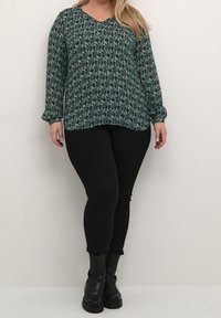Blouse à manches longues en V de couleur vert foncé avec un motif floral complexe ; associée à un jean skinny noir et des bottines noires.