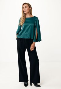 Blusa di raso teal con maniche ampie e spacchi laterali, indossata con pantaloni neri a gamba larga a vita alta e stivaletti neri con tacco.