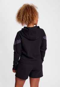 Svart hoodie med huva, med ribbade ärmar och fåll, samt lila detaljer på axlarna. Kombineras med svarta shorts.