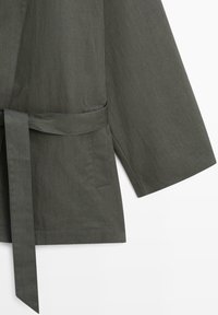 Mouw van een donkergroene olijfgroene jas met een aangehechte stoffen ceintuur en details van zoomnaden op de zak, tegen een witte achtergrond.