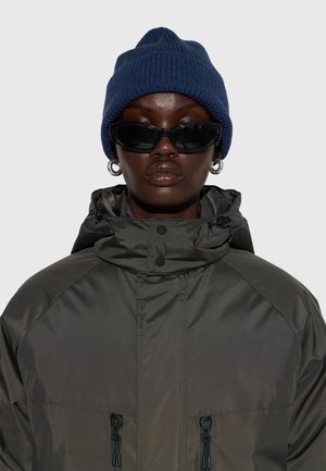 Grüne, mit Reißverschluss versehene, wasserdichte Jacke mit Kapuze, kombiniert mit einer blauen, gestrickten Mütze und schwarzen Sonnenbrillen. Verfügt über strukturierter Stoff und auffällige Nahtakzente.