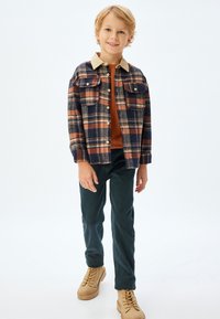 Flanellen shirt met een marineblauwe, oranje en crèmekleurige ruitpatroon, beige kraag, twee borstzakken; gecombineerd met donkergroene broek en beige schoenen.