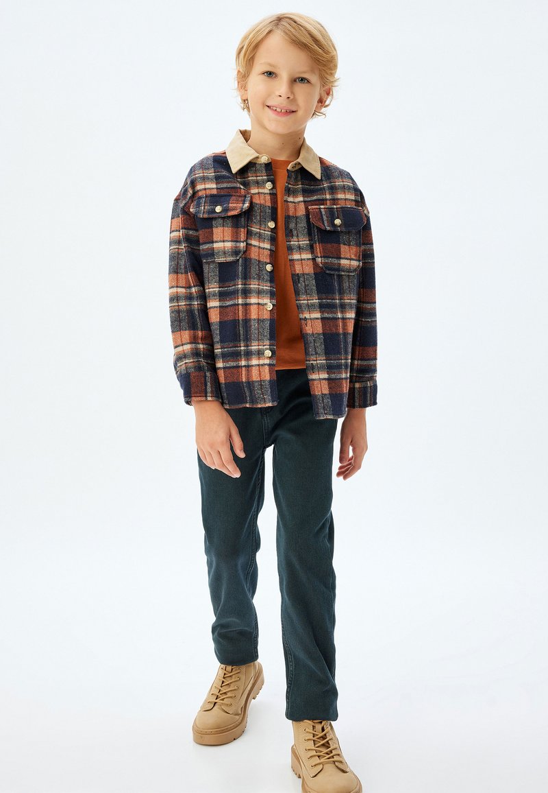 Flanellen shirt met een marineblauwe, oranje en crèmekleurige ruitpatroon, beige kraag, twee borstzakken; gecombineerd met donkergroene broek en beige schoenen.