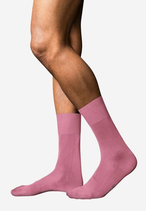 Dos piernas cruzadas con calcetines rosas lisos de caña corta sobre un fondo gris claro.
