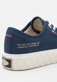 Baskets en toile bleu marine avec des lacets blancs, une semelle en caoutchouc à motif losange et un texte doré "Palladium 77014-FP Canvas 120z Heritage FR" sur le côté.