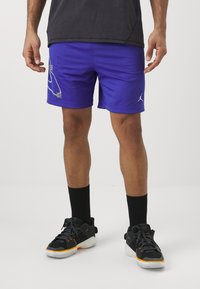 Lilla spordishortsid koos võrkmaterjaliga, millel on valge logo disain. Koos mustade sokkide ja mustade sportjalatsitega, millel on oranžid aktsendid.