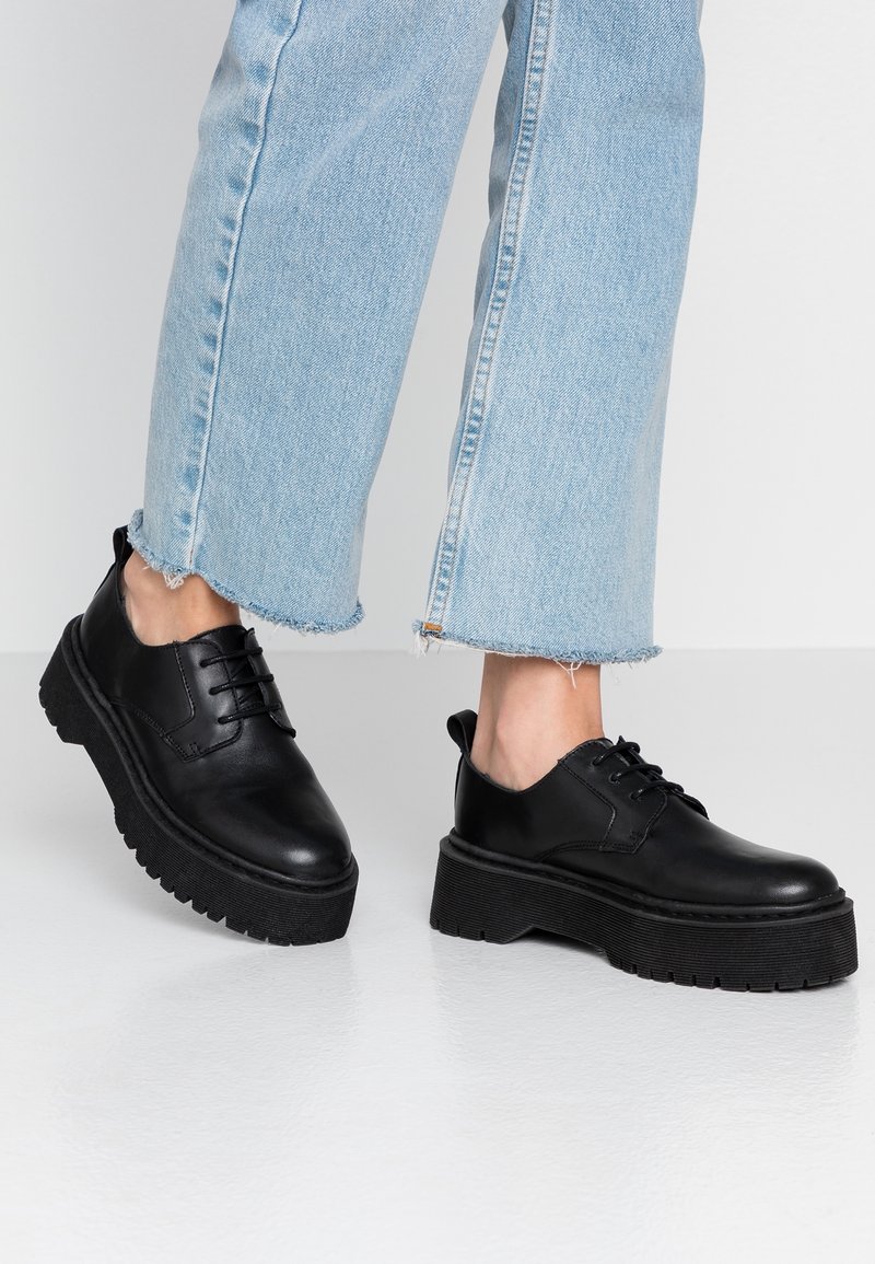 Chaussures à plateforme en cuir noir à lacets portées avec un jean court bleu clair sur fond blanc.