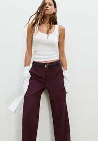 Top in cotone bianco a coste, pantaloni larghi viola scuro con cintura in pelle nera, e una camicia bianca appoggiata su un braccio.