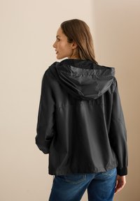 Svart lätt jacka med huva, med en mjuk yta, lös passform och elastiska ärmslut. Baksidan visar ett minimalistiskt designutseende.
