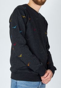 Uomo in un maglione grigio scuro con motivi di uccelli ricamati multicolori, abbinato a jeans azzurri, in piedi con le mani incrociate.