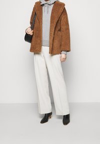 Abrigo de piel sintética marrón con solapas anchas, sudadera gris debajo, pantalones de pierna ancha color crema y botines negros de tacón, sosteniendo un pequeño bolso negro.