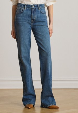 Kvinde iført blå jeans med højtaljet lige ben, hvid skjorte og brune spidse sko, står på et trægulv.