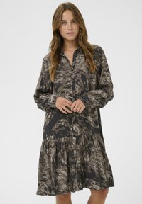 O tânără cu păr lung, purtând o rochie lejeră, cu butoane și un model abstract, având manșete lungi și puffate, pe un fundal simplu.
