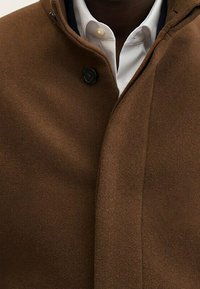 Cappotto di lana marrone con colletto alto, chiusura a singolo bottone e texture liscia. Indossato sopra una camicia bianca, con un design senza cuciture.