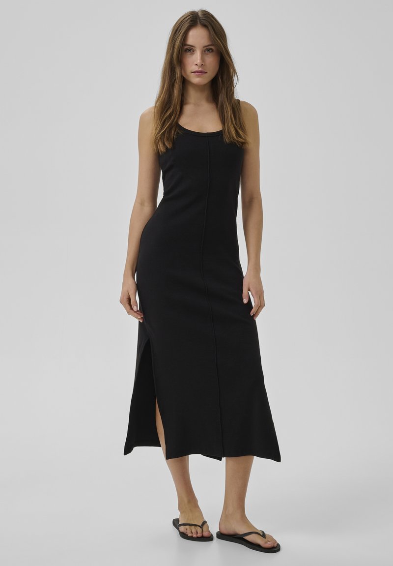 Femme aux longs cheveux bruns portant une robe midi noire sans manches avec une fente sur le côté et des tongs noires, debout devant un fond gris.