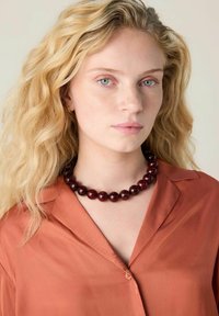 Red semi-precious bead necklace with polished, rounded beads. Model wearing a rust-colored button-up shirt. Wavy blonde hair. Neutral background.