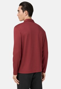 Vyriškis, atsukęs nugarą, vilki ilgomis rankovėmis raudoną marškinių (burgundinę) su apykakle ir juodas kelnes, prieš paprastą baltą foną.