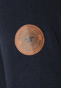 Tissu tricoté bleu marine avec un patch circulaire en cuir beige embossé du texte "PREMIUM QUALITY GOODS" et un motif géométrique à l'intérieur.