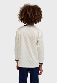 Child with curly hair wearing a long-sleeve white shirt with subtle chevron pattern and navy pants, facing away against a plain background.