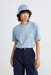 T-shirt bleu clair à manches courtes avec un petit logo jaune sur la poitrine, associé à un pantalon noir marine et un bob en coton côtelé bleu clair.