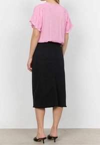 Kvinde iført en pink kortærmet bluse stoppet ned i en højtaljet sort midi-nederdel og sorte hælede mules, stående på et gråt gulv.