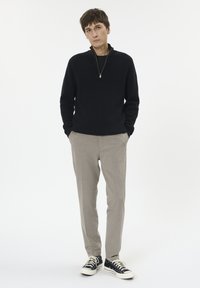 Fekete kötött zipzáras pulóver, szürke nadrág és fekete sportcipő. Laid-back outfit, kényelmes szabással, egyszínű fehér háttér előtt.