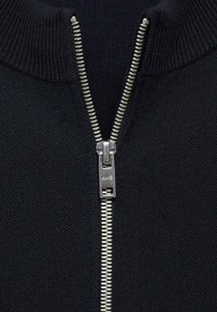 Crna pletena zip-up vesta s visokim okovratnikom, koja ima srebrni metalni patentni zatvarač i rebrastu teksturu duž okovratnika i donjeg ruba.