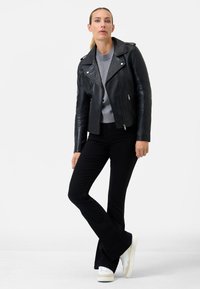 Giacca da motociclista in pelle nera con cerniere argentate, maglione grigio a maglia, jeans neri a campana e sneakers bianche, su uno sfondo semplice.