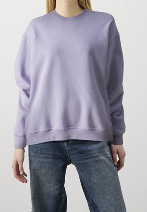 Person iført en ensfarvet lavendel-sweatshirt med rund hals og lange ærmer samt blå jeans, stående mod en hvid baggrund.