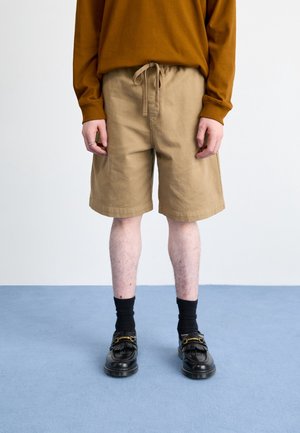 Person trägt beige Shorts mit Kordelzug, schwarze Socken und schwarze Leder-Loafer, steht auf blauem Boden vor weißem Hintergrund.