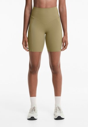 Olivgrüne Radshorts mit hoher Taille, glatter Textur, figurbetontem Design und kontrastierenden Nähten. Kombiniert mit hellgrauen Sportschuhen.