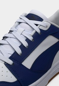 Μπλε και λευκό αθλητικό παπούτσι με υφή δέρματος στην επιφάνεια, που διαθέτει παχύ λευκό πέλμα, λευκά κορδόνια και χρυσή λεπτομέρεια λογότυπου.