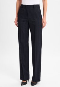 Vrouw in zwarte pinstripe wijdlopende broek met voorzakken en zwarte puntige hakken, staand tegen een witte achtergrond.